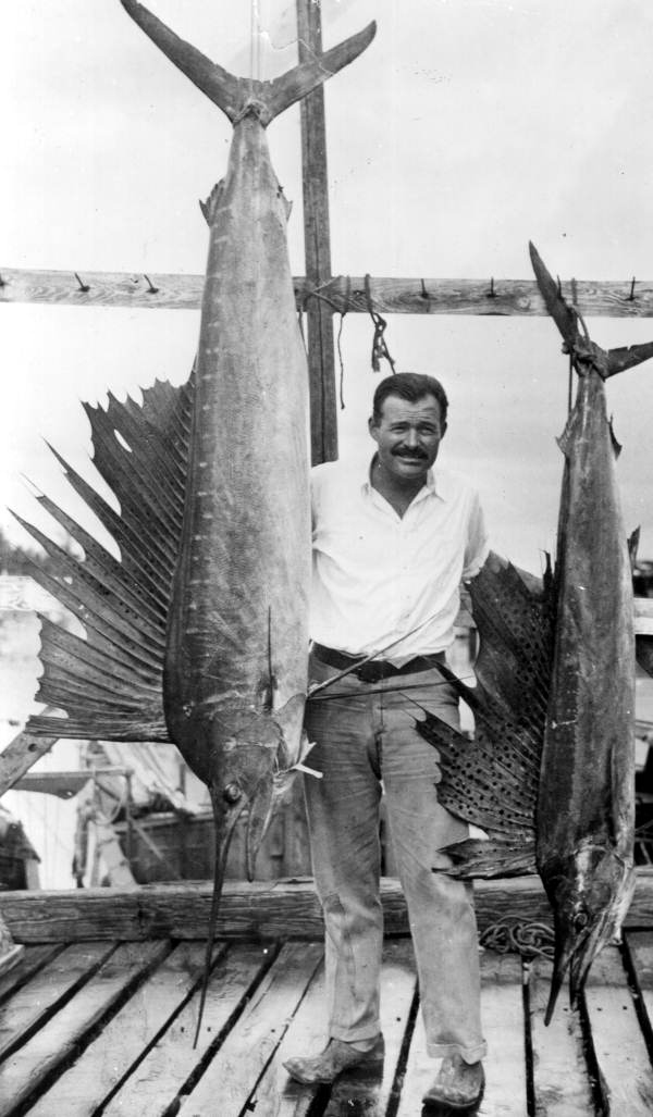 Ernest Hemingway in a white shirt standing on a wooden dock between two large sailfish hanging from a rack in Key West