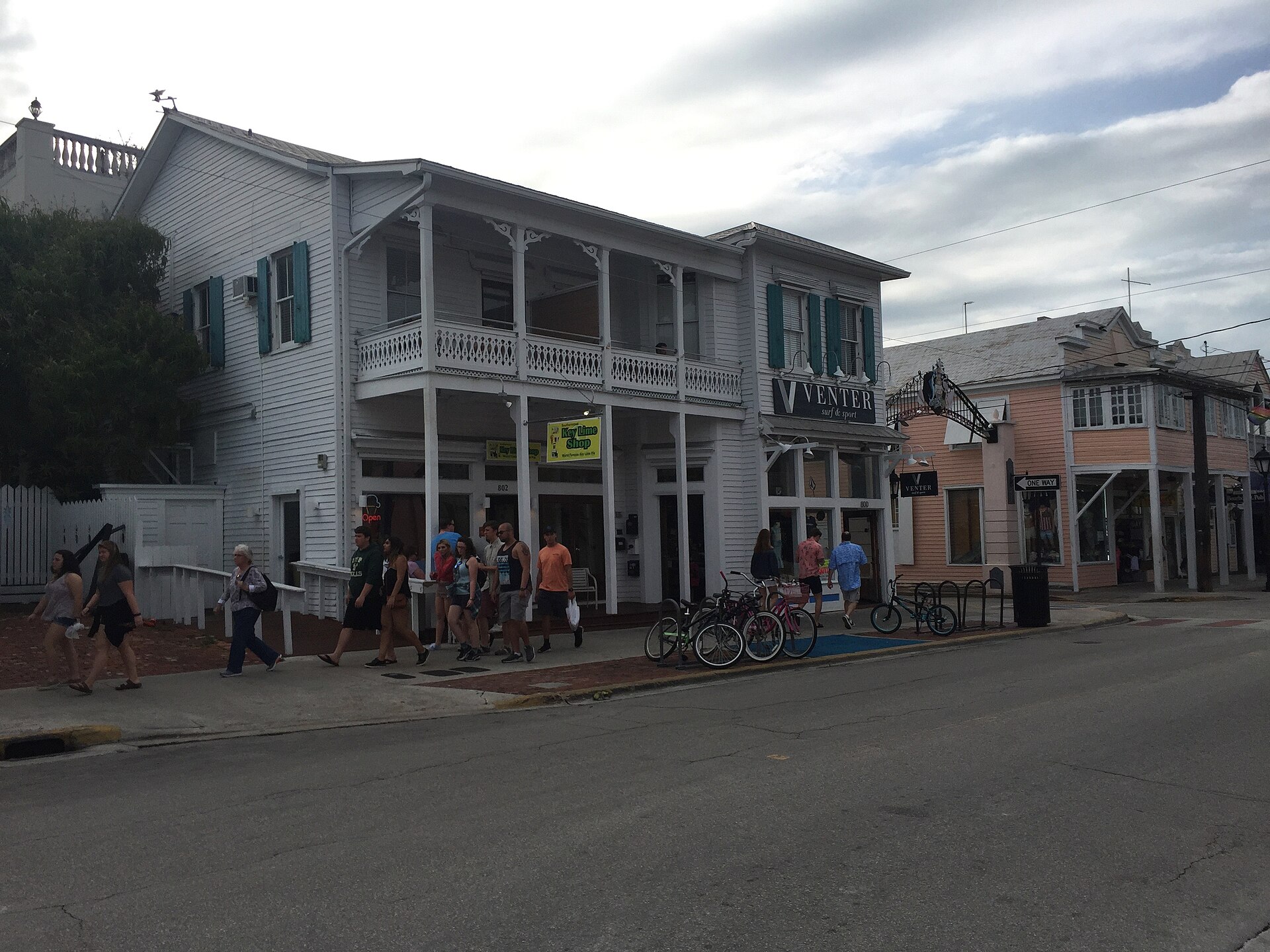 Visitors walking past white wooden buildings with verandas and a key lime pie shop on Duval Street in Key West