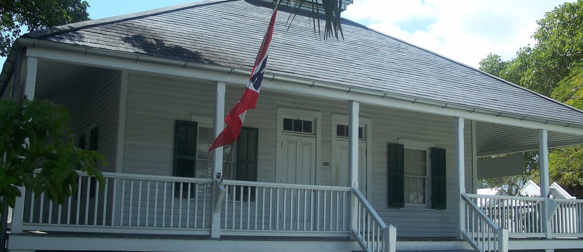 The white wooden Key West Lighthouse Keeper's Quarters with green shutters, a wide porch and a Conch Republic flag under palm fronds