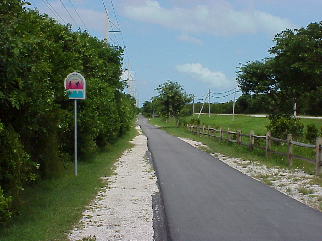 A paved Overseas Heritage Trail path with a trail marker sign running between green shrubs and a wooden fence under blue sky