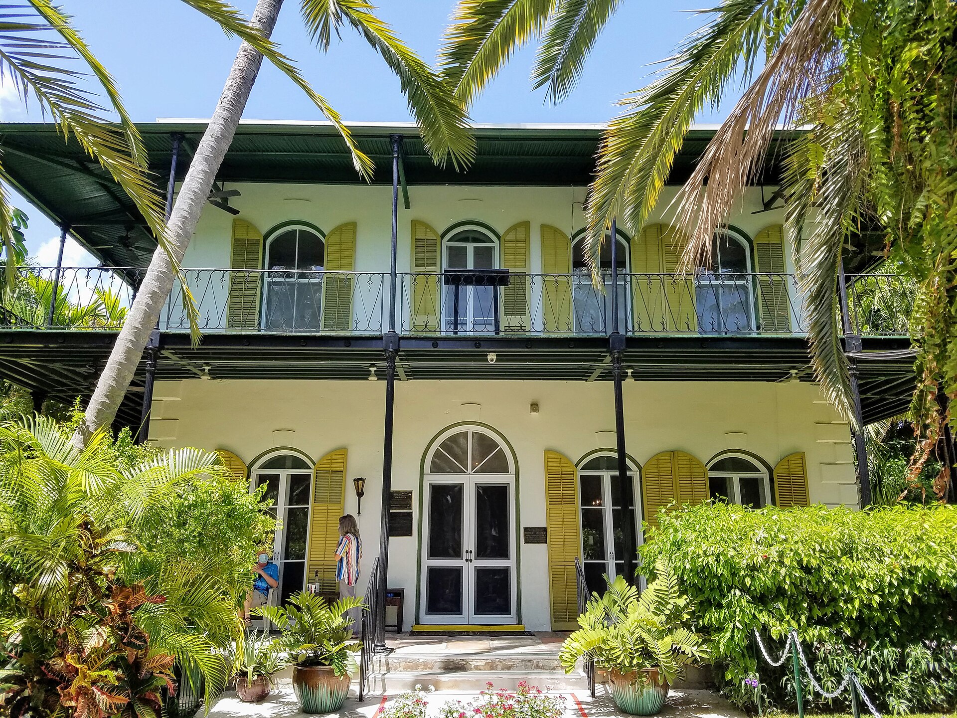The two-story pale yellow Ernest Hemingway Home with arched windows, yellow shutters and a wraparound iron balcony behind palm fronds