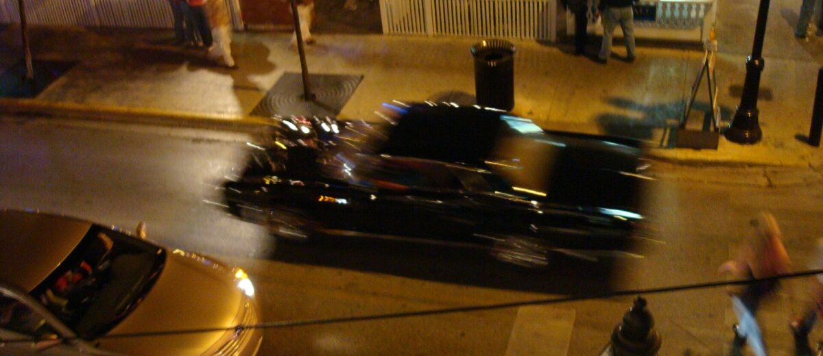 People on a lit sidewalk and outdoor bar terrace on Duval Street at night, with cars passing below