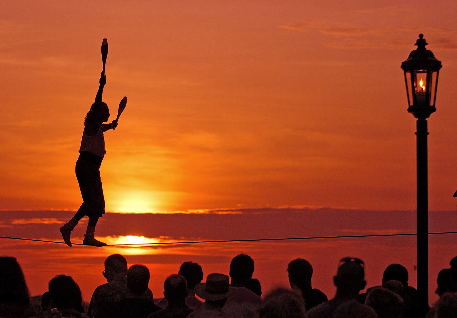A tightrope walker juggling clubs silhouetted against an orange sunset above a crowd, with a street lamp beside them