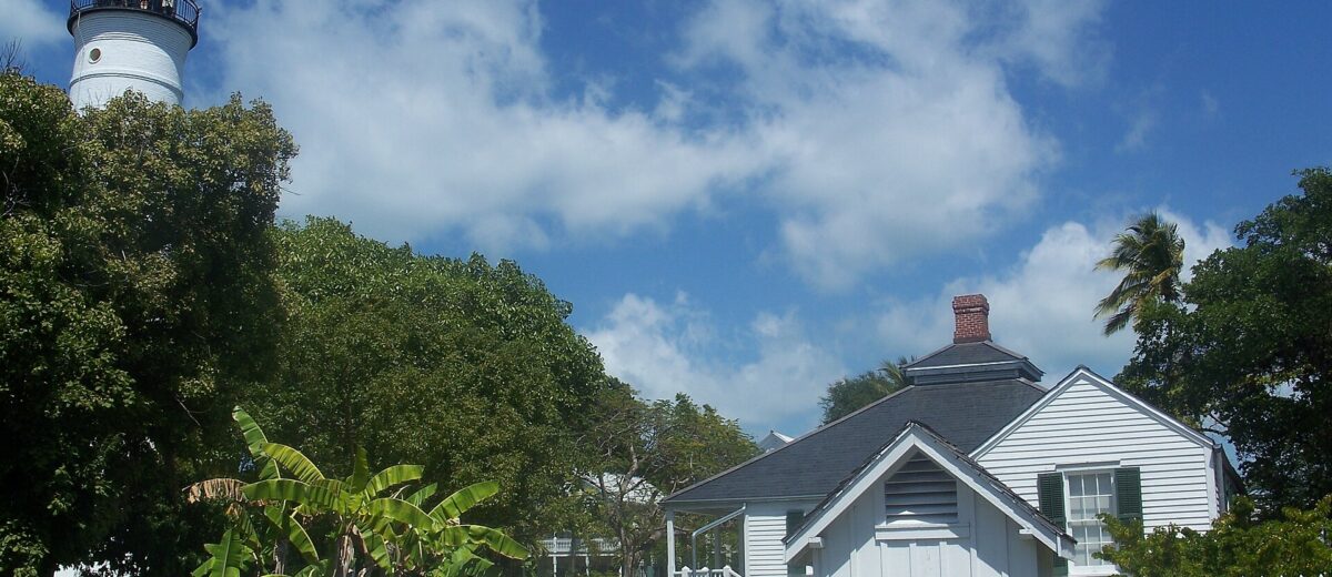 The white Key West Lighthouse rising above trees behind the white wooden keeper's quarters and a white picket fence